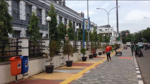 Trotoar ini dihiasi tanaman hias dalam pot warna kuning dan oranye. Ada juga tempat pembuangan sampah di sudut-sudut trotoar.
